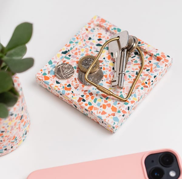 Square catch-all tray with keys and money placed on-top. The tray features a white base with multicoloured terrazzo chips running through. A phone and plant placed next to tray in angled shot.