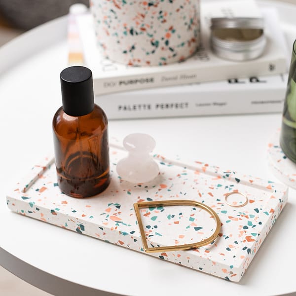 Rectangular trivet tray with terrazzo pattern, with skincare and jewellery resting in tray. The tray is next to a stack of books, plant pots and glass.