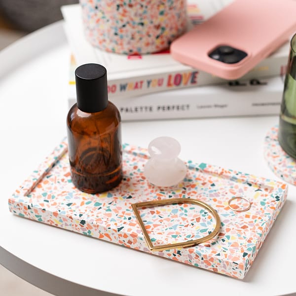 Handmade rectangular decorative tray with colourful terrazzo pattern, with skincare product and jewellery resting in tray. The tray is next to a stack of books, plant pot , phone and glass.