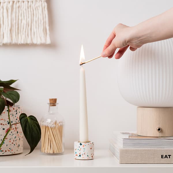 Handmade white candle cup holder with pink, blue and off-white terrazzo chips and brass candle holder insert. Hand in image lighting the dinner candle with a match.