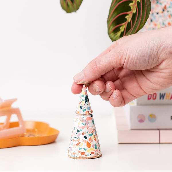Handmade white ring cone with colourful terrazzo pattern with hand placing ring on ring cone. Styled next to a stack of books and decorative tray.