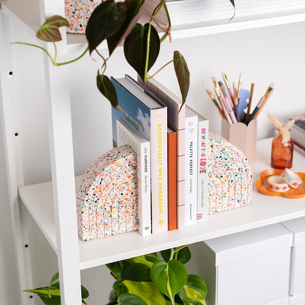 Pair of bookends styled on a shelf, supporting a stack of books. The bookends sit next to a pen pot. The bookends feature a white base with rainbow coloured terrazzo chips running through, in a half arch shape. Each bookend featured an etched half rainbow pattern following the curvature of the bookend. Shot at an angle.