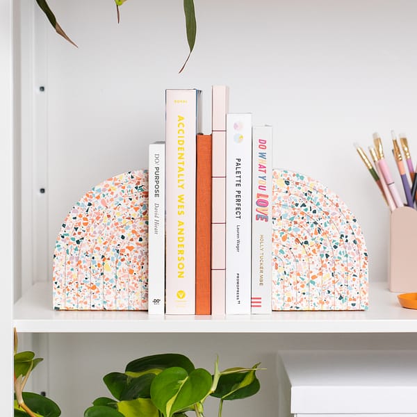 Pair of bookends styled on a shelf, supporting a stack of books. The bookends sit next to a pen pot. The bookends feature a white base with rainbow coloured terrazzo chips running through, in a half arch shape. Each bookend featured an etched half rainbow pattern following the curvature of the bookend.