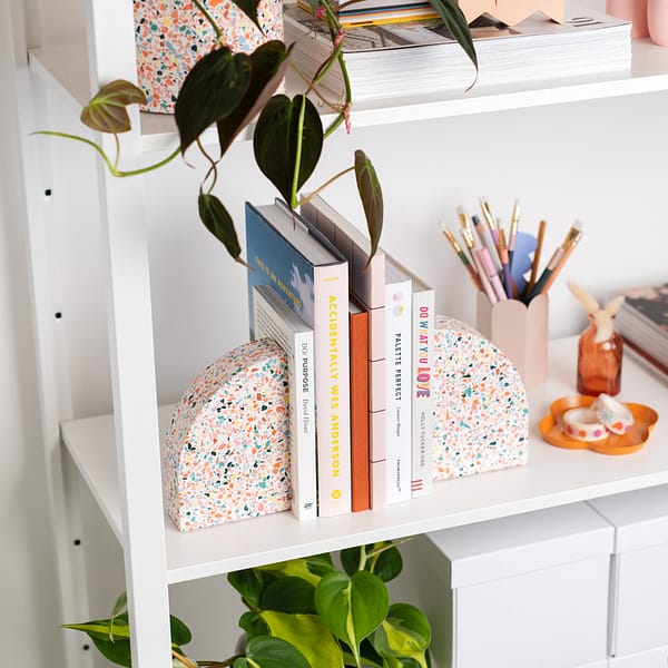 Pair of bookends styled on a shelf, supporting a stack of books. The bookends sit next to a pen pot, trinket tray and a plant pot. The bookends feature a white base with rainbow coloured terrazzo chips running through, in a half arch shape.
