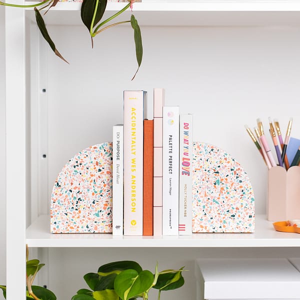 Pair of bookends styled on a shelf, supporting a stack of books. The bookends sit next to a pen pot and are nestled amongst plants. The bookends feature a white base with rainbow coloured terrazzo chips running through, in a half arch shape.