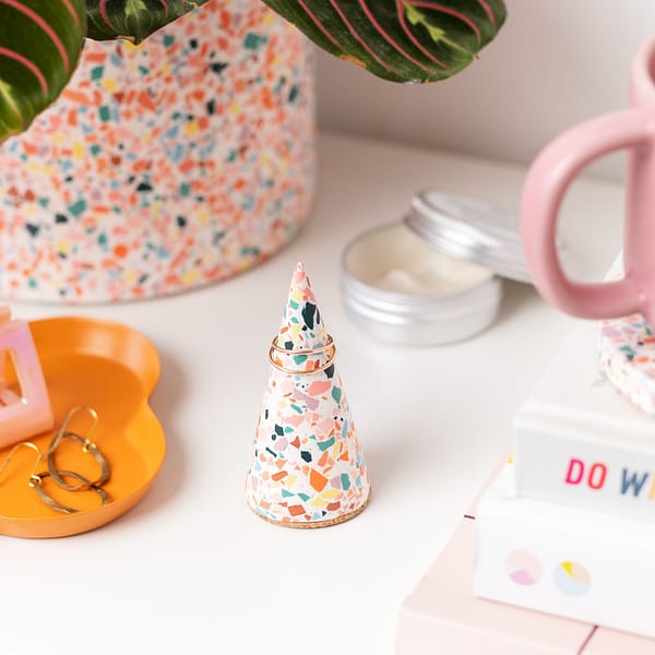 Handmade white ring cone with rainbow terrazzo pattern on table. Styled next to a stack of books, skincare and other colourful home decor items.