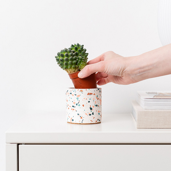 Small white plant pot with terrazzo chips being placed on a desk. Planter is next a to stack of books in a neutral setting.