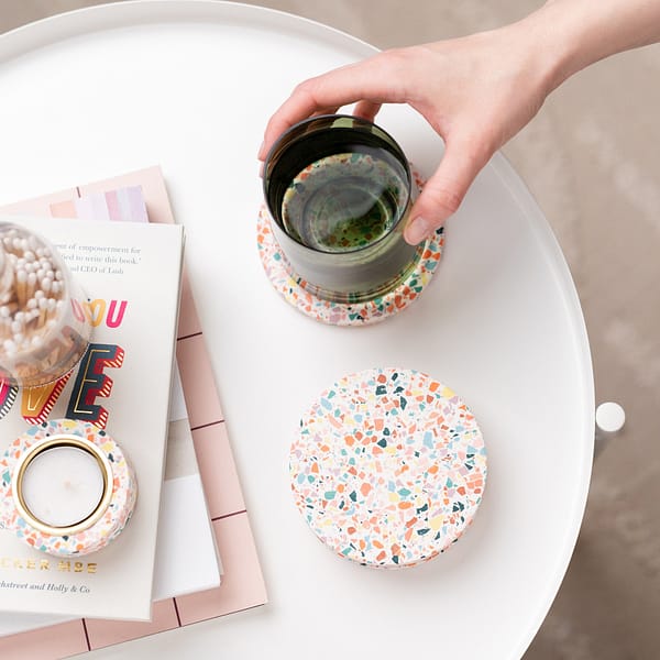 Set of rainbow terrazzo coasters styled on a side table next to a stack of books and terrazzo tealight holder. A hand is placing a glass onto 1 coaster, in a colourfully styled photograph shot top down.