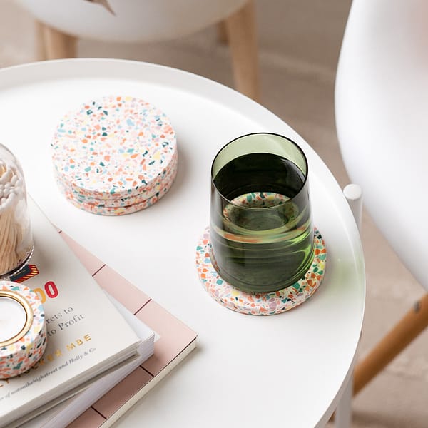 Set of white coasters with a rainbow mosaic-like pattern. The coasters are styled on a side table next to a stack of books and terrazzo tealight holder and matches. A chair is placed nearby.