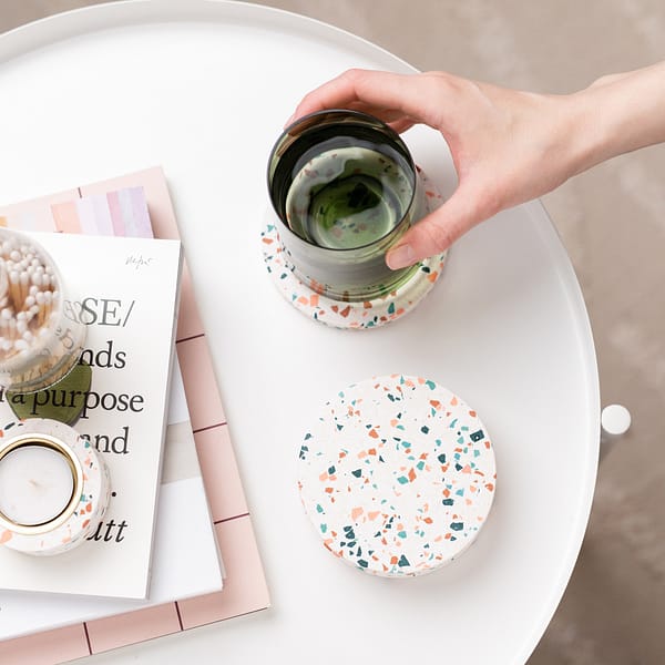 Set of white terrazzo coasters styled on a side table next to a stack of books and terrazzo tealight holder. A hand is placing a glass onto 1 coaster, in a neutrally styled photograph shot top down.