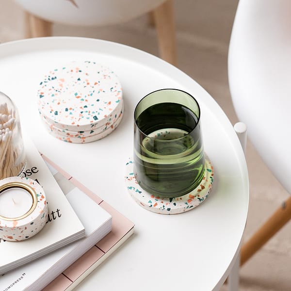 Set of white coasters with a mosaic-like pattern. The coasters are styled on a side table next to a stack of books and terrazzo tealight holder and matches. A chair is placed nearby.