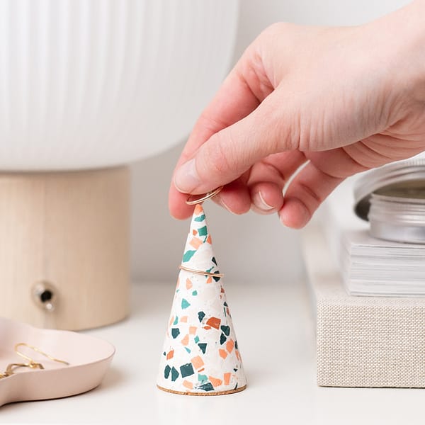 Handmade white ring cone with neutral terrazzo pattern with hand placing ring on ring cone. Styled next to a stack of books and lamps in neutral Scandinavian styled setting.