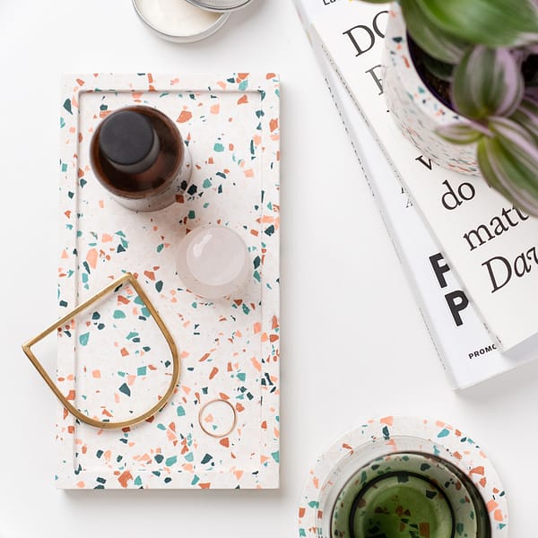 Rectangular decorative tray with terrazzo pattern, with skincare and jewellery resting in tray. The tray is next to a stack of books, coaster and plant and is shot as a flatlay.