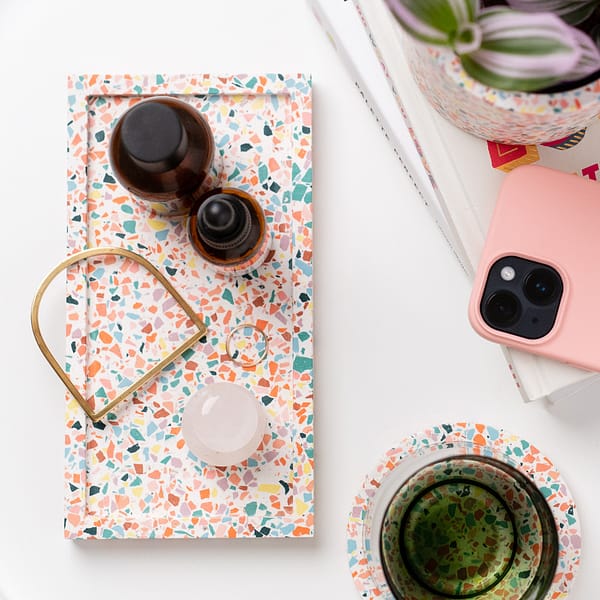 Rectangular display tray with colourful handmade terrazzo pattern, with skincare and jewellery resting in tray. The tray is next to a stack of books, coaster, plant and phone and is shot as a flatlay.