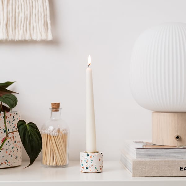 Single dinner candle, which is lit, sitting in a white terrazzo candle holder. The holder is styled on a table next to books, table lamp, matches and a plant. All items have a scandi neutral style.