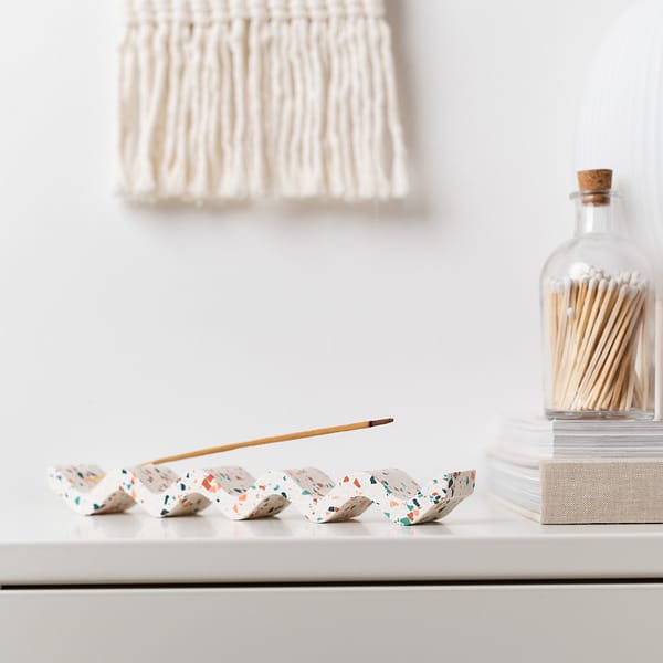 Incense holder with a wavy design and featuring a white base and pink, blue and off-white terrazzo chips. Styled on a cabinet with incense stick and next to books and matches in a scandi styled home.