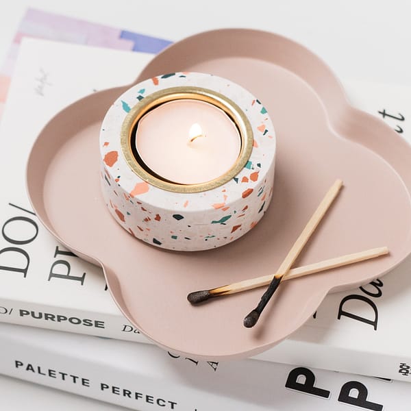 Single tealight holder with a tealight inside sitting on a trinket tray. The tealight holder has a white base with pink, white and off-white terrazzo patterning and a brass tealight insert. Close up image set on a stack of books and on a trivet tray next to used matches.