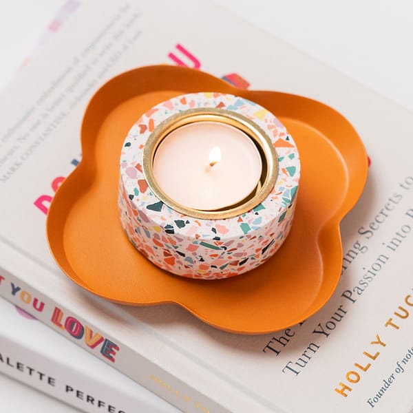 Single tealight holder with a tealight inside sitting a trinket tray. The tealight holder has a white based with coloured terrazzo patterning. Close up image set on a stack of books.