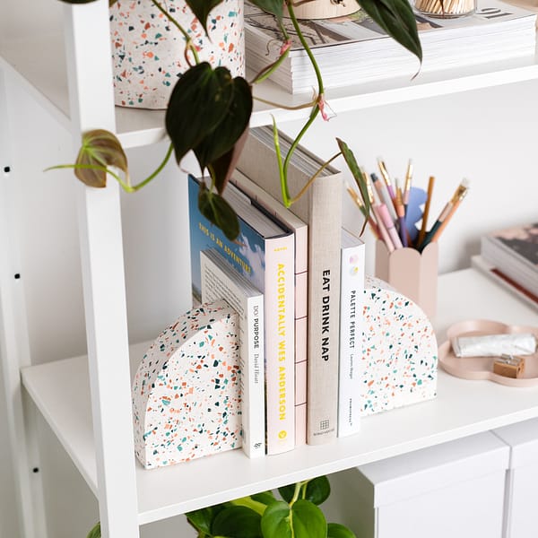 Pair of bookends styled on a shelf, supporting a stack of books. The bookends sit next to a pen pot, trinket tray and a plant pot. The bookends feature a white base with pink, blue, dark blue and off-white coloured terrazzo chips running through, in a half arch shape.