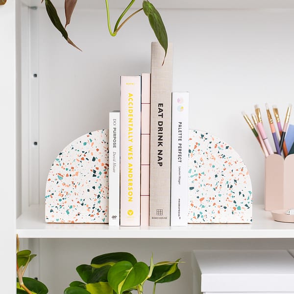 Pair of bookends styled on a shelf, supporting a stack of books. The bookends sit next to a pen pot and are nestled amongst plants. The bookends feature a white base with pink, blue, teal and off-white coloured terrazzo chips running through, in a half arch shape.