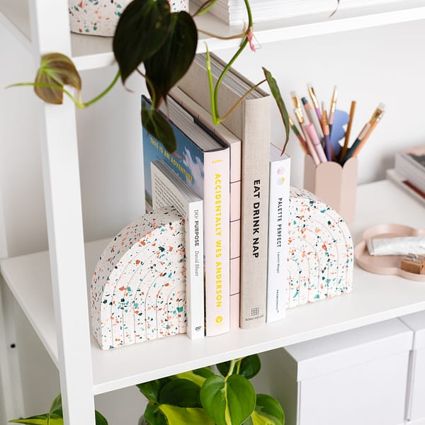Pair of bookends styled on a shelf, supporting a stack of books. The bookends sit next to a pen pot. The bookends feature a white base with pink, blue and off-white coloured terrazzo chips running through, in a half arch shape. Each bookend featured an etched half rainbow pattern following the curvature of the bookend. Shot at an angle.