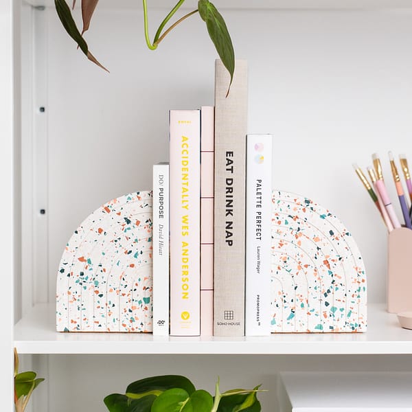 Pair of bookends styled on a shelf, supporting a stack of books in a neutral colourway. The bookends sit next to a pen pot. The bookends feature a white base with blue, pink and neutral coloured terrazzo chips running through, in a half arch shape. Each bookend featured an etched half rainbow pattern following the curvature of the bookend.