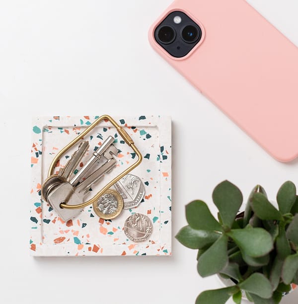 Square catch-all tray with keys and money placed on-top. The tray features a white base with pink, blue and off-white terrazzo chips running through. A phone and plant placed next to tray in a flatlay shot.
