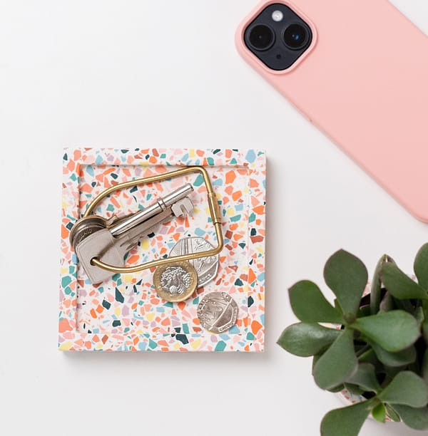 Square catch-all tray with keys and money placed on-top. The tray features a white base with multicoloured terrazzo chips running through. A phone and plant placed next to tray in a flatlay shot.